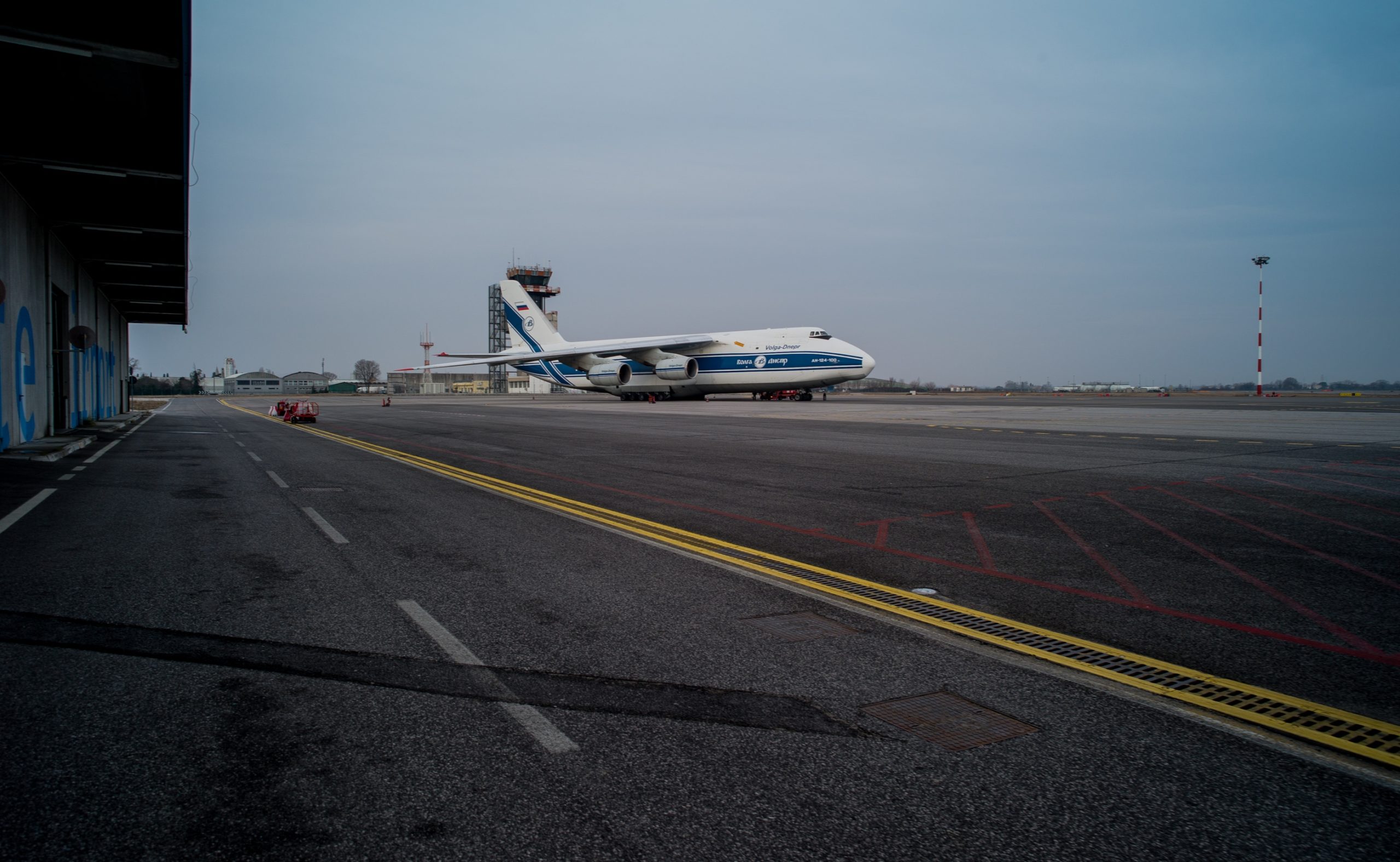 A large cargo aircraft parked on the tarmac at Trieste Airport, with the airport control tower and surrounding buildings visible in the background